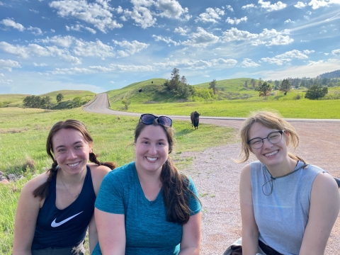 three people smile together with rolling green hills, blue skies, and bison in the background. 