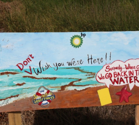 A protest sign at the entrance to Grand Isle, LA. The sign is handpainted and reads "Don't wish you were here! Seriously, when can we go back in the water?"