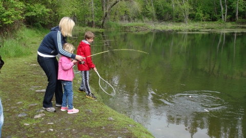 An adult helps a young girl reel in a fish while another child helps net the fish. They are standing on the edge of the water in a green natural setting.
