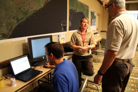 Three people are busy at work in the Operations Center during the aftermath of the Deepwater Horizon oil spill. One person is focused on a computer screen, with a map of the impacted area over their head. The two other people are talking to each other. 