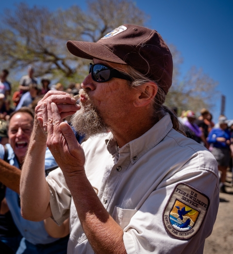 A man wearing a U.S. Fish and Wildlife Service uniform kissing a razorback sucker. Large crowd in background.