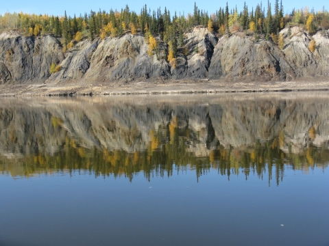 A bluff with trees on top reflects in the still water of a river.