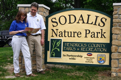 A man and woman stand together reading a brochure next to a park sign.