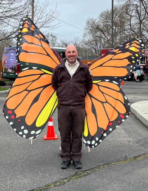 A man wearing brown clothes is standing in front of a orange and black butterfly kite. 