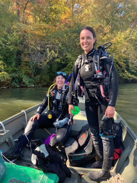 Two people smile from inside a boat, wearing diving gear and equipment.