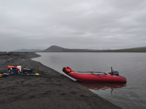 A red boat sits near the black sand shores of Mapsorak lagoon; grey hills under a grey sky emerge in the distance. A few supplies, including a white bucket, blue bin, and orange life vest sit on the sand near the boat. 