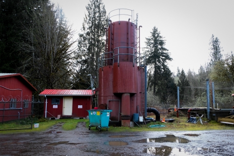 A red, two-story, cylindrical metal building stands to the right of a small, similarly red, square shack. A turquoise dumpster rests in front of the cylindrical building. To the right, a net is strung above a square, man-made pond which is bordered by metal sides and pipes. In the foreground, puddles gather in a muddy gravel road, reflecting the grey sky. 