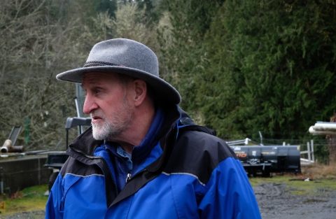 Man stands in profile, facing left. He is visible from his chest and up. He wears a felt grey trail hat and two electric blue windbreakers. Lush, green trees dominate the landscape behind him. Metal storage equipment sits on the gravel, puddled ground behind him.