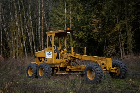 A bright yellow motor grader stands in front of a deep green forest backdrop. On the side of the grader is a white sticker depicting red and black fish artwork, that says: “Nisqually Public Works Department.”