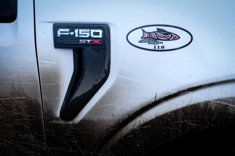 A close-up of a side of a white Ford pickup truck, just above the front right wheel. The truck’s metal “F-150 STX” letters are printed on a black, comma-shaped piece of the door. A sticker to the right of this text portrays a red and black Nisqually drawing of a salmon. The fish is contained within an oval, much wider than it is tall. The numbers 118 printed beneath the fish. The entire truck is wet with rain. 