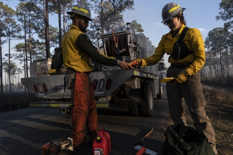 Two firefighters stand in front of a fire engine and hand each other supplies needed to cut down trees in a recent burn area. They are standing on a paved road and you can see burned trees and vegetation in the background.