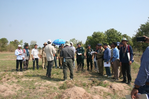 Twenty or so people stand outdoors in a semi-circle around two people giving instructions