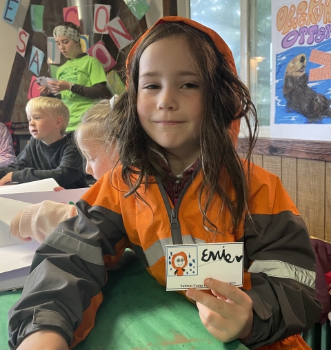 a girl in an orange coat holds a handmade fishing license with a drawing of herself on it