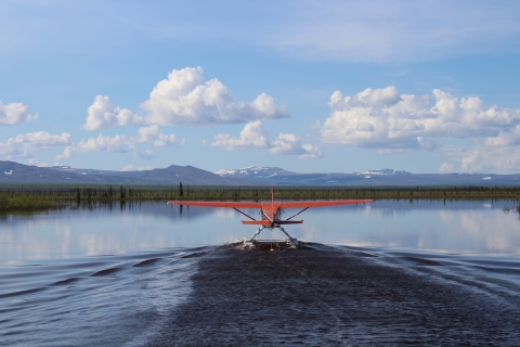 A red floatplane takes off from a lake and heads toward mountains.