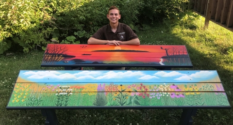 A man smiles while standing in front of two painted benches. One features a wetland scene with a sun setting over the water and trees on either side. The other features a variety of flowers in a field with blue skies and clouds over them.