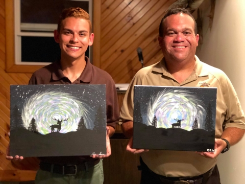 Two people hold identical paintings featuring a silhouette of a deer in front of a starry night sky.