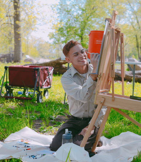Logan Sauer paints on an easel out in nature on a sunny day. 