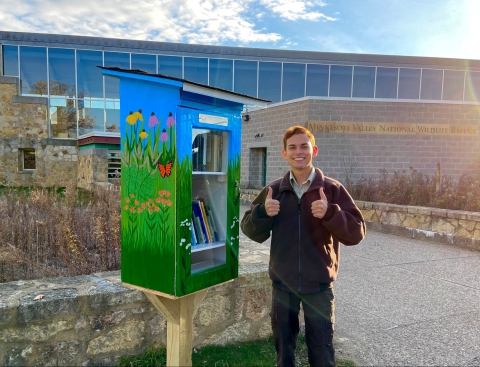 A man holds his thumbs up while standing next to a tiny library painted with grass, butterflies and flowers.