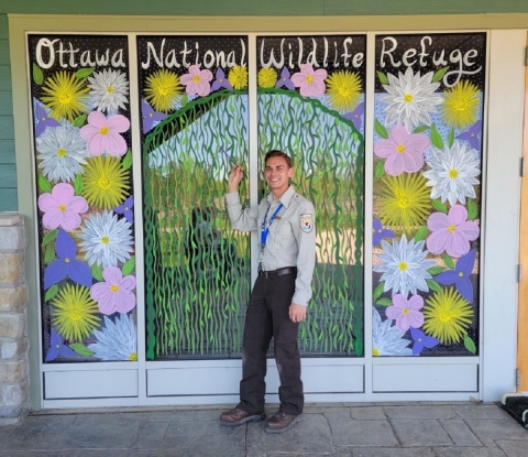 A person holds a paintbrush next to a window mural featuring various flowers on vegetation. The mural reads "Ottawa National Wildlife Refuge"