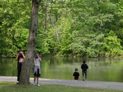 four children stand by keney park pond watching. Two face the camera using binoculars, and two are turned away towards the water
