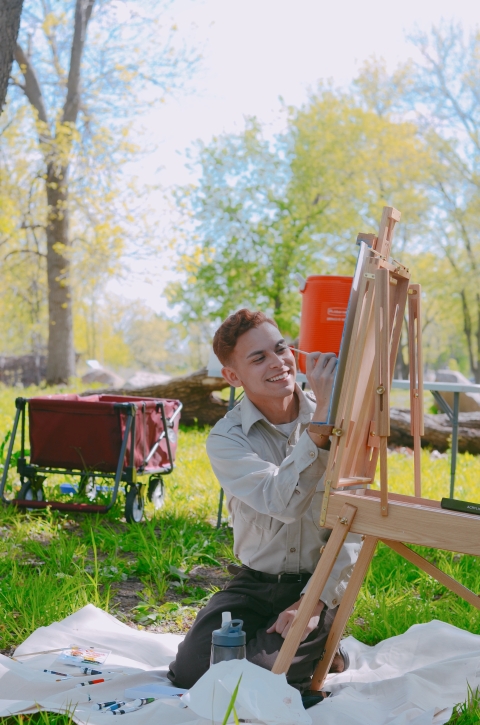 a man sits on a sheet outside in a city park and paints on an easel. 