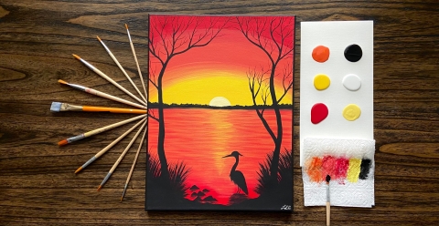A set of paintbrushes, paints on paper, and a painting of a sun setting over a wetland with a bird and trees in the foreground
