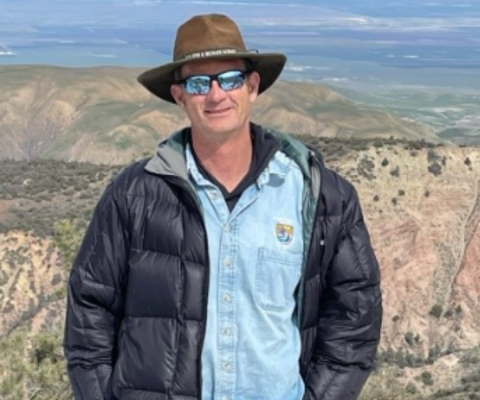Jeff Phillips stands in front of a mountainous landscape. He is wearing a USFWS logo'd shirt, a jacket, sunglasses, and a wide brimmed hat. 