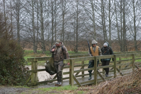 Three people dressed in camouflage gear with one person carrying a hunting rifle as they cross a wooden bridge in a forested wetland