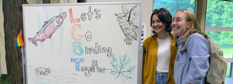 Two young women grin for a camera next to a "Let's Go Birding Together" sign on a whiteboard, with a small rainbow flag attached to the left side of the board.