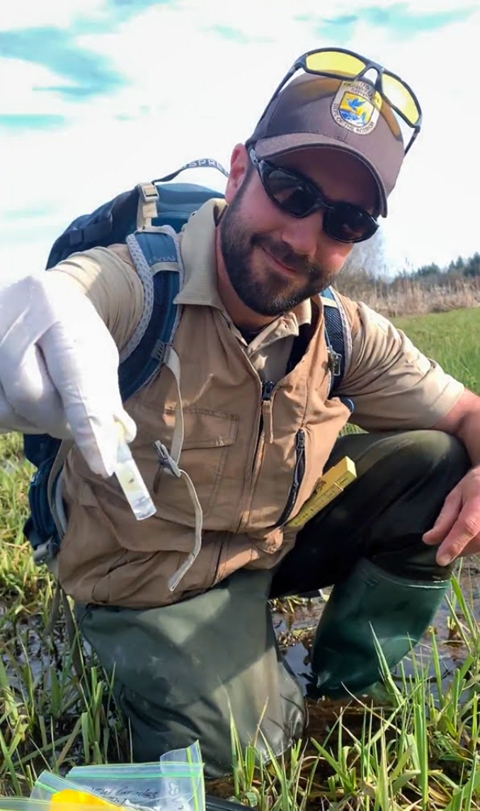 A smiling man with black beard and dark sunglasses, wearing waders and a Service hat, kneels in a shallow wetland and hold a vial out toward the camera in a gloved hand.