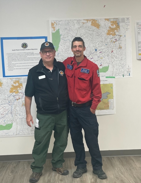 Bill Waln stands next to Dallas Gatchell, Wildfire Technician for Dawson Creek Fire Zone, BC Wildfire Service. Both individuals are in their team gear and are standing in front of a wall with maps on it.
