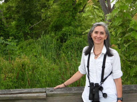 A smiling women with binoculars around her neck wearing a white shirt with greenery in the background