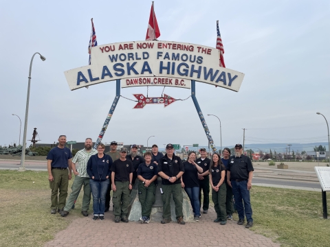 A group of firefighters pose in front of a sign that says "You are Now Entering the World Famous Alaska Highway. Dawson Creek, B.C." There are 14 people total.