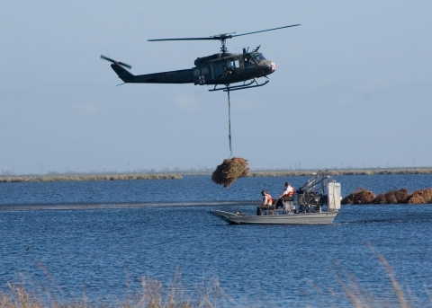Two people stand on an airboat in a body of water as a helicopter hovers nearby 