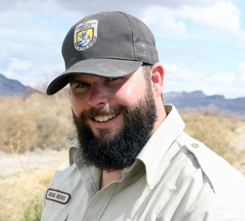 Man in baseball cap with U.S. Fish and Wildlife Service shield with a black beard smiles with blue mountains in backdrop.
