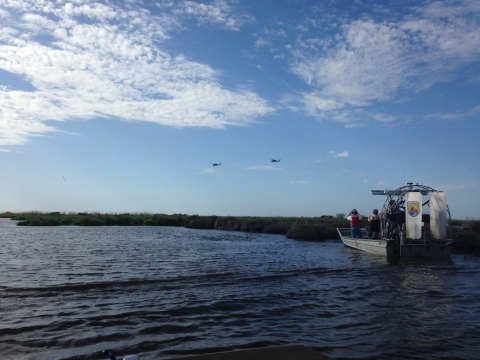 Two people stand on an airboat in a body of water as two helicopters fly in the distance