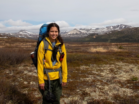 A women wearing a backpack stands infront of a snow speckled mountain range and tundra.
