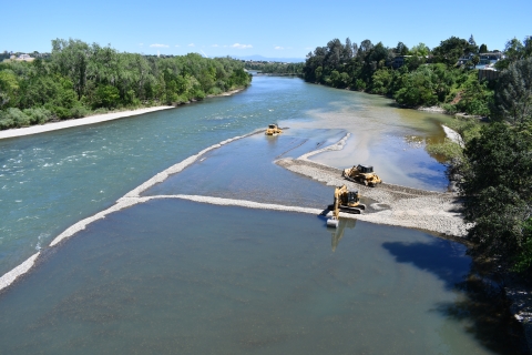 Aerial view of a river with two bull dozers and an excavator working on spreading gravel along the bank of the river. 