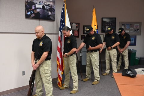 Five honor guard members stand in a line. The first and last are holding rifles and the three in the middle are holding flags. Each has they head down in honor.