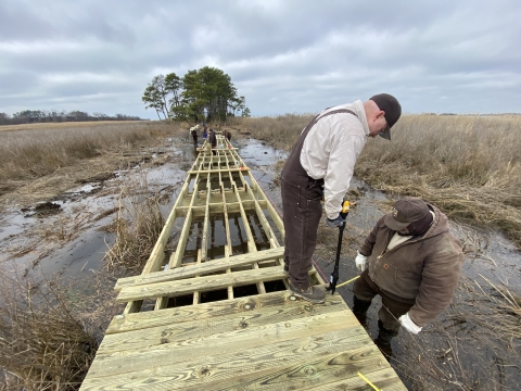 A worker in foreground stands on top of an unfinished boardwalk that stretches into distance, while another in foreground stands alongside it.