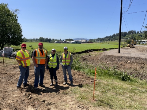 Four people stand side by side in a dirt and grassy area wearing bright safety vests and hats.