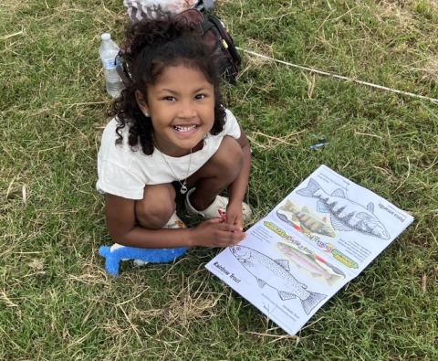 A child looking at the camera smiling and showing off a trout being colored in a coloring book. 
