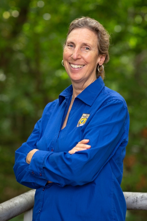 Director Williams stands outside, wearing a blue shirt with Service logo. She is smiling and her arms are folded. The wooded grounds of the National Conservation Training Center are in the background.