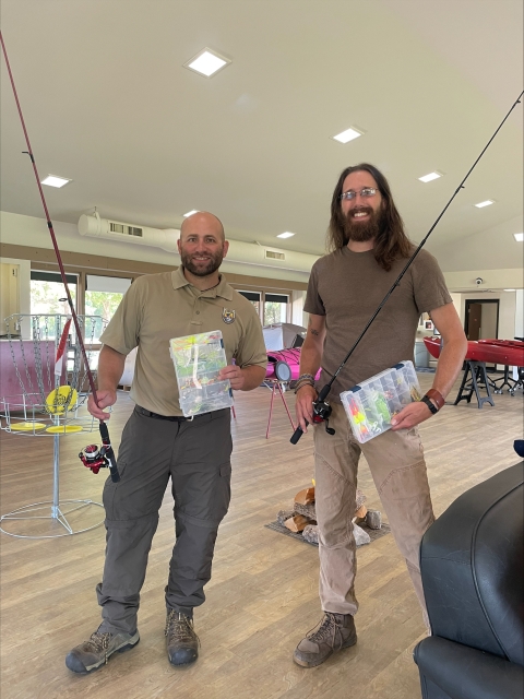 Two individuals stand in a room holding fishing poles and tackle boxes.