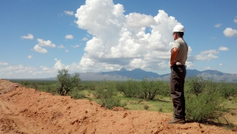 A man in a hard hat stands on a mound of dirt and looks out over patchy grass and tall bushes with mountains in the background.