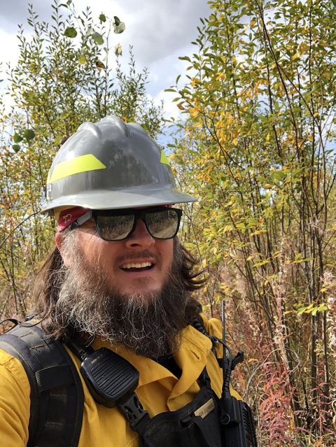 A smiling man in a hard hat, sunglasses, and yellow wildland fireman shirt stands in front of trees.
