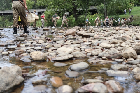 Students in chest waders and carrying nets walk toward a river bank