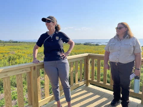 A woman in a black shirt and gray pants and a woman in a Fish and Wildlife Service uniform stand on a wooden platform above green and yellow vegetation