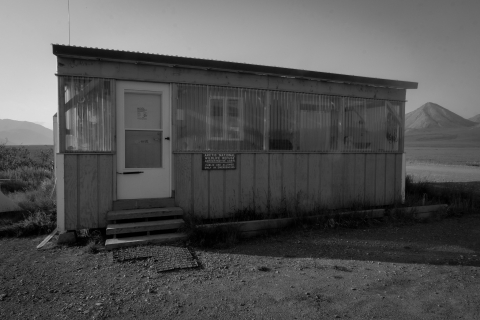 A black and white image of the U.S. Fish and Wildlife Service's bunkhouse at the Arctic National Wildlife Refuge. The rectangular bunkhouse, with its wooden front facade and plastic porch windows, sits at the end of an airstrip near the Galbraith Lake campground. In the background, the Brooks Range stands.