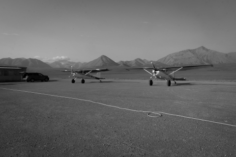 A black and white photo of two bush planes sit side by side on a gravely airstrip near Galbraith Lake campground, just outside the Arctic National Wildlife Refuge. A chord runs along the ground in the foreground, out of shot to the left, where an SUV is parked in front of a rectangular wooden bunkhouse. In the background, the Brooks Range stands.